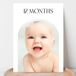 以下が含まれることがあります： 上部に「12ヶ月」の文字が入った写真プリント。画像には、泡まみれで笑顔の赤ちゃんが写っています。プリントは木製のトレイに飾られています。