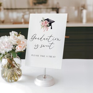 May include: A white graduation sign with the text "Graduation is sweet" and "PLEASE TAKE A TREAT!" is displayed on a silver stand. The sign is decorated with a graduation cap and floral accents. A vase of white and pink flowers sits to the left.