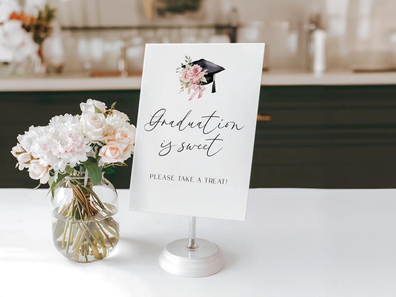 May include: A white graduation sign with the text "Graduation is sweet" and "PLEASE TAKE A TREAT!" is displayed on a silver stand. The sign is decorated with a graduation cap and floral accents. A vase of white and pink flowers sits to the left.