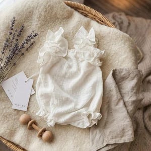 Ivory baby girl clothes displayed flat on a cozy blanket next to a wooden rattle and dried lavender. This breathable premium cotton newborn gift is an elegant, thoughtful choice for a baby shower gift or baptism celebration.