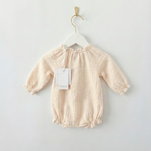 Lylou long-sleeve baby romper hanging against a white wall. Showcases the functional smocked elastic neckline for easy dressing. Crafted from 100% breathable vanilla cotton for newborn baby girl clothes.