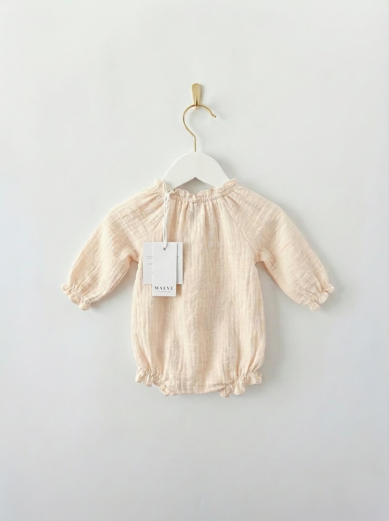 Lylou long-sleeve baby romper hanging against a white wall. Showcases the functional smocked elastic neckline for easy dressing. Crafted from 100% breathable vanilla cotton for newborn baby girl clothes.
