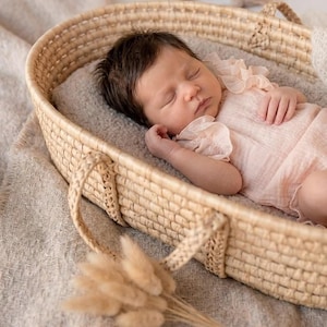 Sleeping newborn girl in a woven Moses basket wearing a creamy vanilla ruffle romper. Soft cotton fabric flatters delicate skin. Heartwarming lifestyle portrait and emotive newborn photography setup with warm, neutral earth tones.