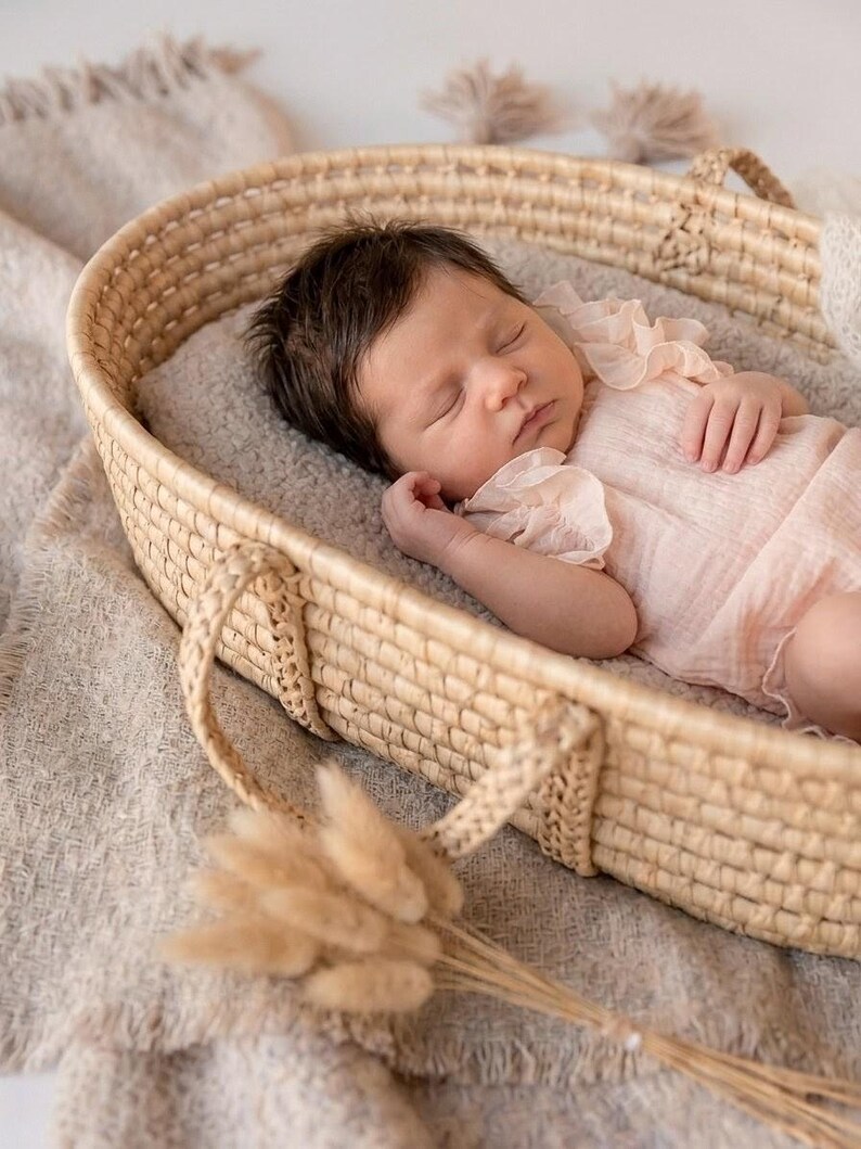 Sleeping newborn girl in a woven Moses basket wearing a creamy vanilla ruffle romper. Soft cotton fabric flatters delicate skin. Heartwarming lifestyle portrait and emotive newborn photography setup with warm, neutral earth tones.