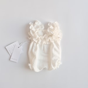 Back-facing flat lay of an ivory baby romper displaying its symmetrical smocked silhouette and ruffled shoulder details. A minimalist, camera-ready baby shower gift designed for timeless studio portraits.