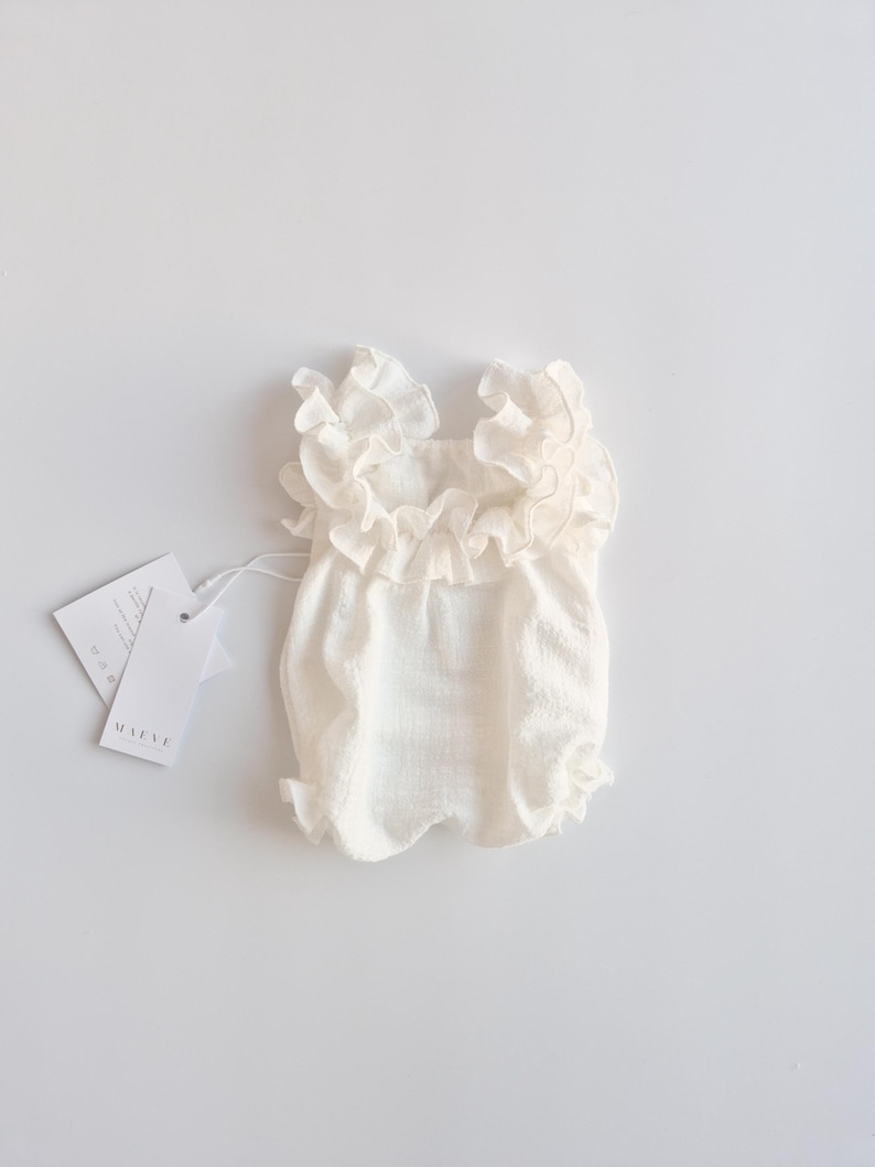 Back-facing flat lay of an ivory baby romper displaying its symmetrical smocked silhouette and ruffled shoulder details. A minimalist, camera-ready baby shower gift designed for timeless studio portraits.