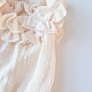 Extreme close-up of textured cotton fabric on a newborn girl romper. Creamy vanilla ruffled collar showcasing the delicate weave and soft, breathable drape. High-quality studio foundation for heirloom baby portraits.