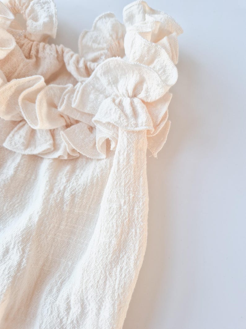 Extreme close-up of textured cotton fabric on a newborn girl romper. Creamy vanilla ruffled collar showcasing the delicate weave and soft, breathable drape. High-quality studio foundation for heirloom baby portraits.