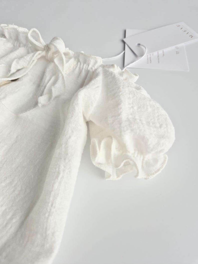 Detailed macro shot of an ivory infant outfit highlighting the dainty puff sleeve and soft woven texture. A perfect zero-prep newborn photo prop, capturing romantic simplicity and earthy elegance for professional lifestyle galleries.
