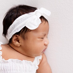 Close-up profile portrait of a sleeping baby girl. Highlighting the textured white gauze cotton knot bow headband and the ruffled strap of the smocked top. Natural window light creates a soft, timeless aesthetic for this sweet baby girl gift.