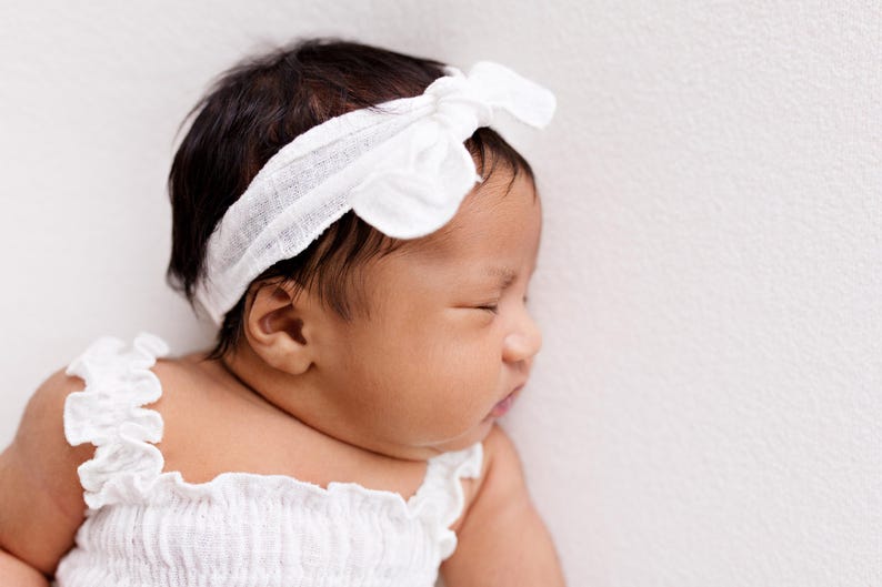 Close-up profile portrait of a sleeping baby girl. Highlighting the textured white gauze cotton knot bow headband and the ruffled strap of the smocked top. Natural window light creates a soft, timeless aesthetic for this sweet baby girl gift.
