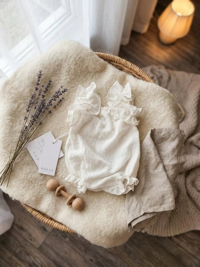 Ivory baby girl clothes displayed flat on a cozy blanket next to a wooden rattle and dried lavender. This breathable premium cotton newborn gift is an elegant, thoughtful choice for a baby shower gift or baptism celebration.