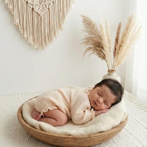 Sleeping newborn in a rustic wooden bowl with macrame and pampas grass backdrop. Wearing the vintage-style Lylou vanilla romper. Perfect boho aesthetic newborn outfit girl for studio fine art photography.