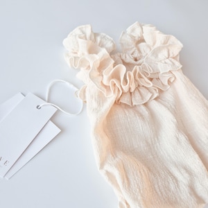 High-stretch newborn romper in creamy vanilla hue. Premium textured cotton fabric with intricate frilled shoulder details. Timeless newborn girl clothes perfect for natural light photography and cohesive maternal styling.