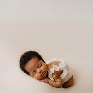 Sleeping infant wearing an ivory off-white newborn outfit, posed on a soft neutral backdrop. Close-up highlights the 3D hand-pleated back ruffles. A classic minimalist baby romper perfect for timeless, low-friction photography.