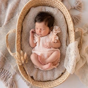 Sleeping newborn girl inside a rustic woven basket wearing a creamy textured baby romper. The cotton fabric and ruffle shoulders create a soft, ethereal vibe, serving as highly sought-after newborn photography props for studio sessions.
