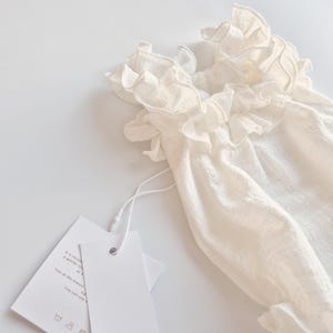 Close-up of smocked baby clothes detailing a hand-pleated ruffle neckline. The breathable ivory cotton weave shows deep textural shadows, highlighting the premium fabric quality perfect for a baptism gift girl.