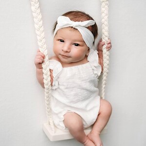 Awake newborn baby sitting on a macrame rope swing wearing an ivory ruffle baby romper and matching bow headband. Soft studio lighting highlights the textured cotton fabric, showcasing an ideal newborn photo props styling.