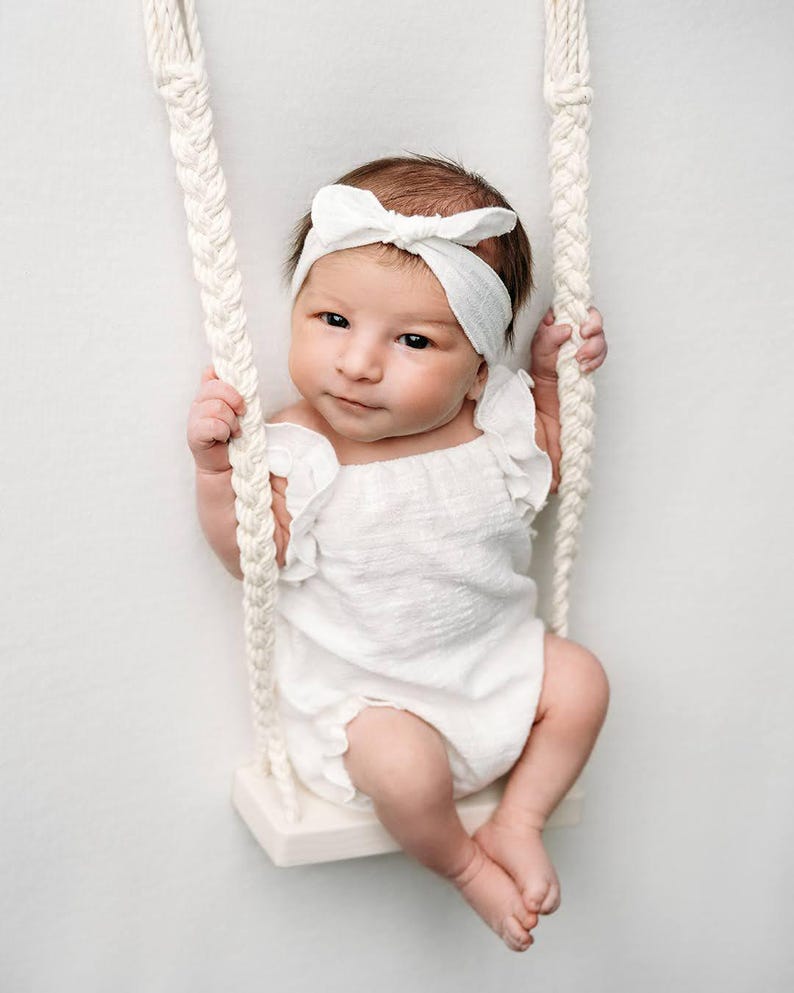 Awake newborn baby sitting on a macrame rope swing wearing an ivory ruffle baby romper and matching bow headband. Soft studio lighting highlights the textured cotton fabric, showcasing an ideal newborn photo props styling.