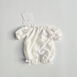 Top-down view of a minimalist white crinkle cotton newborn romper isolated on a seamless background. The dainty puff sleeves and delicate silhouette present an earthy, vintage-inspired studio essential for fine art galleries