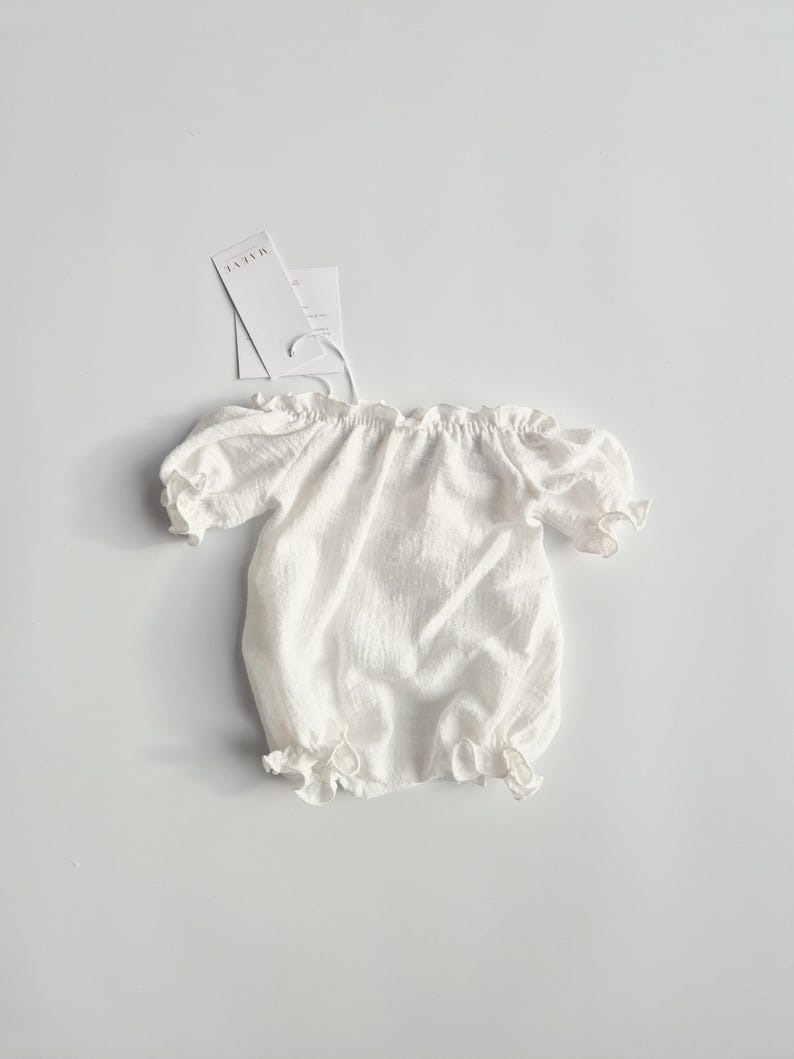 Top-down view of a minimalist white crinkle cotton newborn romper isolated on a seamless background. The dainty puff sleeves and delicate silhouette present an earthy, vintage-inspired studio essential for fine art galleries