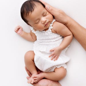 May include: A newborn infant with dark hair sleeping peacefully while wearing a white sleeveless romper. The garment features a smocked bodice with a ruffled neckline and a ruffled waistline, crafted from a lightweight, textured fabric.