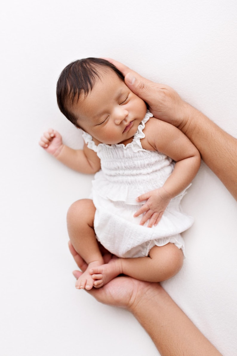 May include: A newborn infant with dark hair sleeping peacefully while wearing a white sleeveless romper. The garment features a smocked bodice with a ruffled neckline and a ruffled waistline, crafted from a lightweight, textured fabric.