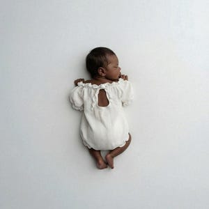 Newborn sleeping on its stomach against a minimalist white studio backdrop, revealing an elegant tie-back keyhole detail on the ivory romper. This functional design is a staple for authentic, low-key newborn photography sessions.