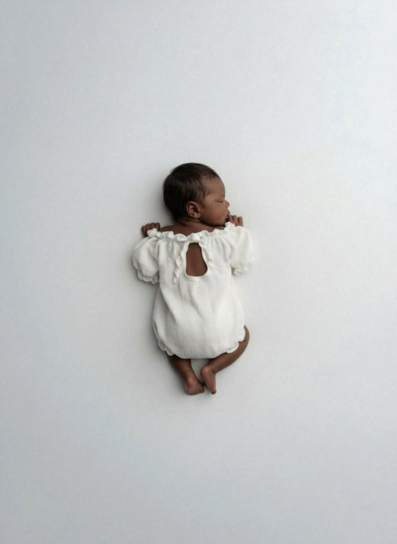 Newborn sleeping on its stomach against a minimalist white studio backdrop, revealing an elegant tie-back keyhole detail on the ivory romper. This functional design is a staple for authentic, low-key newborn photography sessions.
