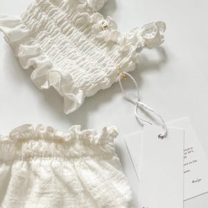 Crisp flat lay shot of a white crinkled cotton two-piece infant outfit featuring elegant paper brand tags attached with a gold pin. The bright, high-key studio lighting emphasizes the boutique presentation, ideal for premium newborn gifts.