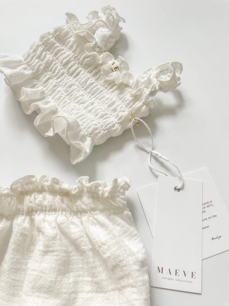 Crisp flat lay shot of a white crinkled cotton two-piece infant outfit featuring elegant paper brand tags attached with a gold pin. The bright, high-key studio lighting emphasizes the boutique presentation, ideal for premium newborn gifts.