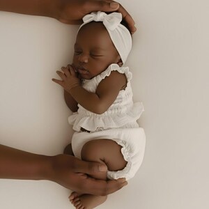 Photographer's hands carefully support a sleeping newborn wearing a white two-piece baby romper. The studio strobe lighting emphasizes the tactile, soft cotton puffy bloomers and smocked top, showcasing versatile and functional newborn props.