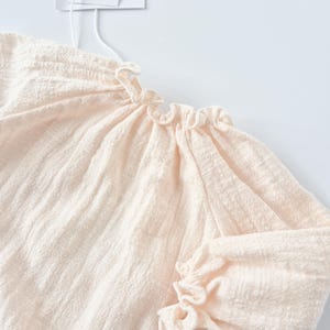 Macro view of the Lylou newborn outfit focusing on the delicate smocked elastic ruffled cuffs and neckline. The pre-softened warm vanilla fabric provides a gentle, sensory-friendly fit for sensitive skin.