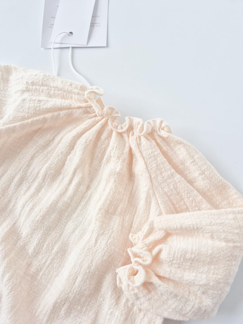 Macro view of the Lylou newborn outfit focusing on the delicate smocked elastic ruffled cuffs and neckline. The pre-softened warm vanilla fabric provides a gentle, sensory-friendly fit for sensitive skin.