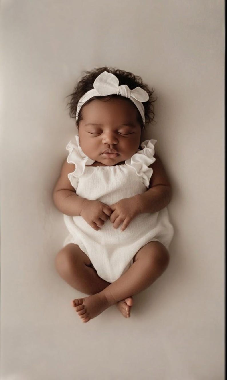 Frontal view of a sleeping infant in a luminous off-white newborn romper and coordinating bow. Showcases the secure smocked bodice and elasticized leg openings designed to conceal diapers seamlessly during studio portrait sessions.