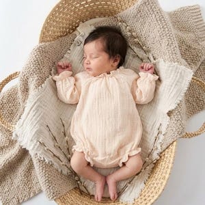 Newborn girl peacefully sleeping in a woven basket. Wearing the Lylou warm vanilla crinkled cotton long-sleeve romper. Earthy, ethereal newborn photography styling with natural pussy willow accents.