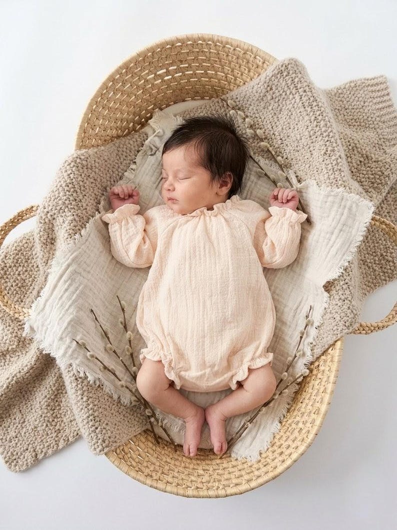 Newborn girl peacefully sleeping in a woven basket. Wearing the Lylou warm vanilla crinkled cotton long-sleeve romper. Earthy, ethereal newborn photography styling with natural pussy willow accents.