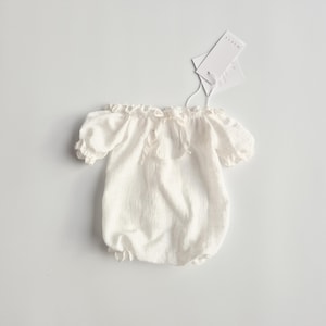 Flat lay of a white studio wardrobe baby outfit showcasing an adjustable 2-in-1 elastic neckline. Made of unsteamed crinkle cotton, these versatile newborn photo props transition smoothly from boatneck to off-the-shoulder styles.