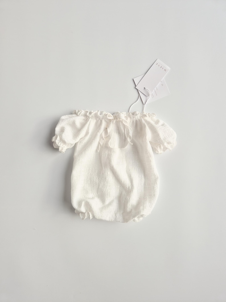 Flat lay of a white studio wardrobe baby outfit showcasing an adjustable 2-in-1 elastic neckline. Made of unsteamed crinkle cotton, these versatile newborn photo props transition smoothly from boatneck to off-the-shoulder styles.