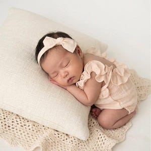 Peaceful newborn girl resting on a cream cushion, dressed in a warm vanilla newborn outfit. She wears a coordinated soft-bow matching headband and ruffle baby romper, capturing a heartwarming, earthy aesthetic for hospital announcements.