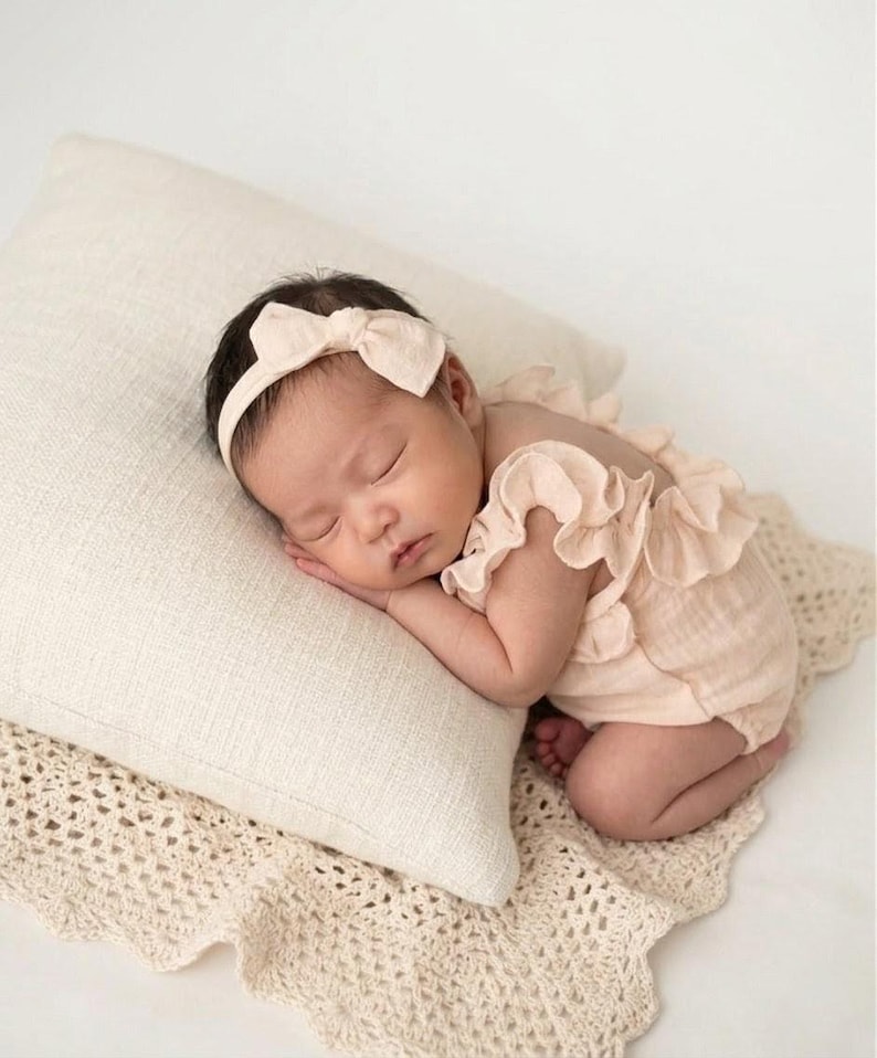 Peaceful newborn girl resting on a cream cushion, dressed in a warm vanilla newborn outfit. She wears a coordinated soft-bow matching headband and ruffle baby romper, capturing a heartwarming, earthy aesthetic for hospital announcements.