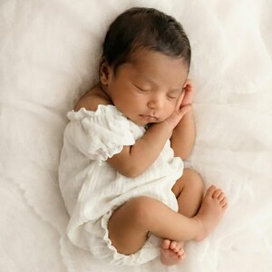 Close up of an infant wearing a textured white cotton smocked romper styled off-the-shoulder. The ethereal, minimalist vibe makes these boho baby clothes perfect for highlighting delicate newborn features in a natural light studio.