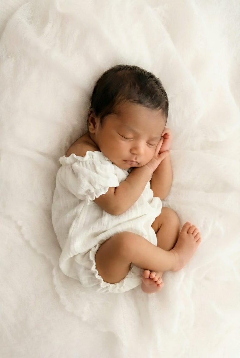 Close up of an infant wearing a textured white cotton smocked romper styled off-the-shoulder. The ethereal, minimalist vibe makes these boho baby clothes perfect for highlighting delicate newborn features in a natural light studio.