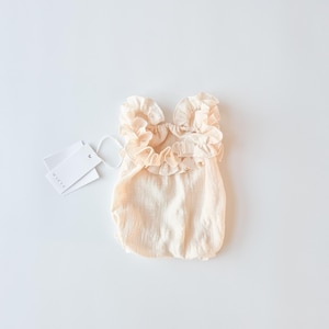 Creamy vanilla newborn girl outfit flat lay. Breathable textured cotton baby romper with oversized frill collar and brand tags. Elegant baby shower gift or hospital announcement wardrobe staple in a soft, bright studio setting.