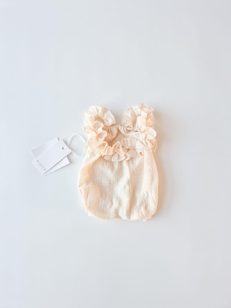 Creamy vanilla newborn girl outfit flat lay. Breathable textured cotton baby romper with oversized frill collar and brand tags. Elegant baby shower gift or hospital announcement wardrobe staple in a soft, bright studio setting.