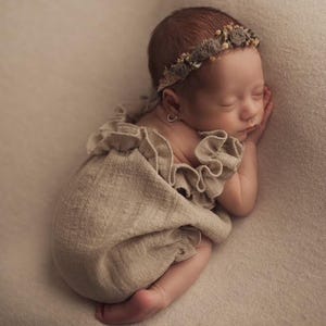 Op de afbeelding: Een beige, getextureerde romper met ruches op de schouderbandjes en een bijpassende bloemenhaarband met kleine stoffen bloemetjes. Het kledingstuk is gemaakt van een zachte, geweven stof en is ontworpen voor een baby, met een neutrale uitstraling.