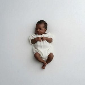 Infant sleeping soundly on a seamless white backdrop, dressed in an ivory off-the-shoulder smocked romper. The pure, romantic simplicity of this breathable baby hospital outfit doubles flawlessly as an elegant baptism outfit girl.