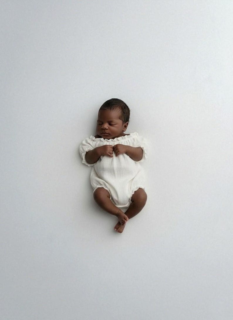 Infant sleeping soundly on a seamless white backdrop, dressed in an ivory off-the-shoulder smocked romper. The pure, romantic simplicity of this breathable baby hospital outfit doubles flawlessly as an elegant baptism outfit girl.