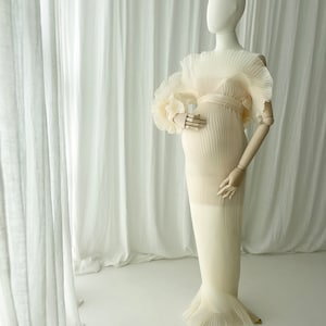 Full-length view of the Celessia gown in warm apricot on a mannequin. The sheer pleated fabric and voluminous sleeves are displayed against sheer white curtains, emphasizing the gown's ethereal flow and structure.
