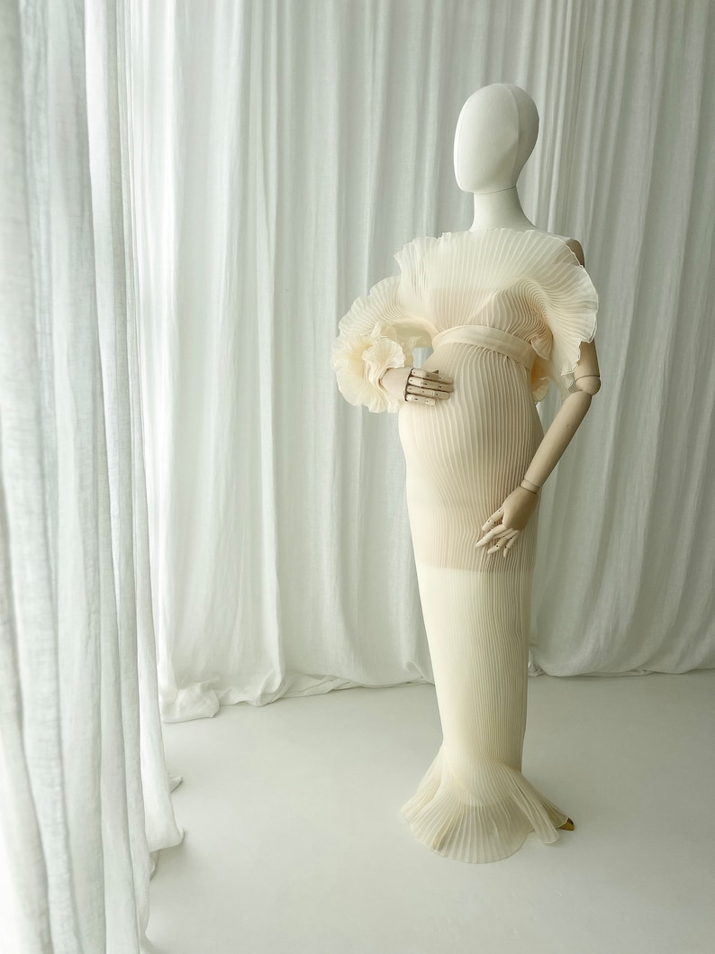 Full-length view of the Celessia gown in warm apricot on a mannequin. The sheer pleated fabric and voluminous sleeves are displayed against sheer white curtains, emphasizing the gown's ethereal flow and structure.